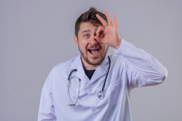 Доктор молодого человека нося белое пальто и стетоскоп делая одобренный знак с рукой и пальцы смотря через знак с большой улыбкой