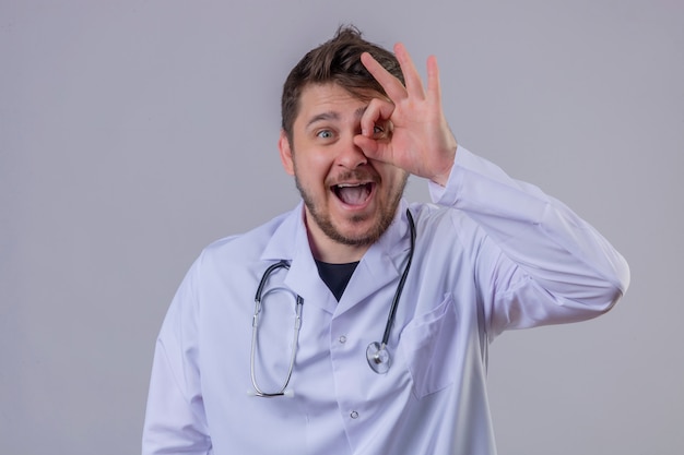 Young man doctor wearing white coat and stethoscope doing ok sign with hand and fingers looking through sign with big smile