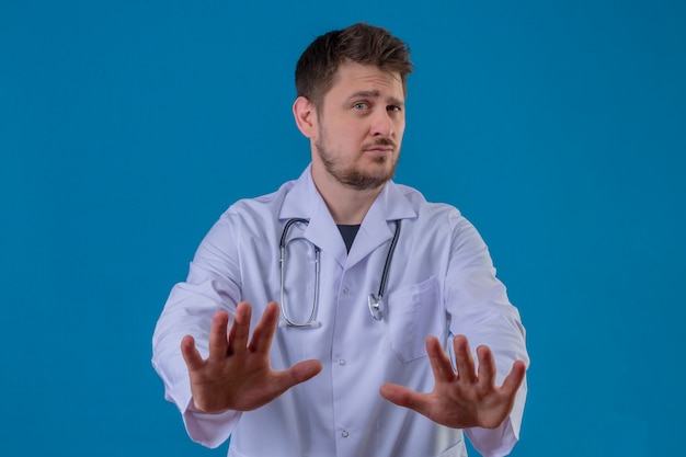 Young man doctor wearing white coat and stethoscope doing stop sign, defense gesture over isolated blue background