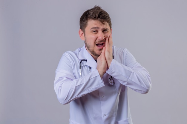 Доктор молодого человека нося белое пальто и стетоскоп чувствуя боль страдая от сильной зубной боли