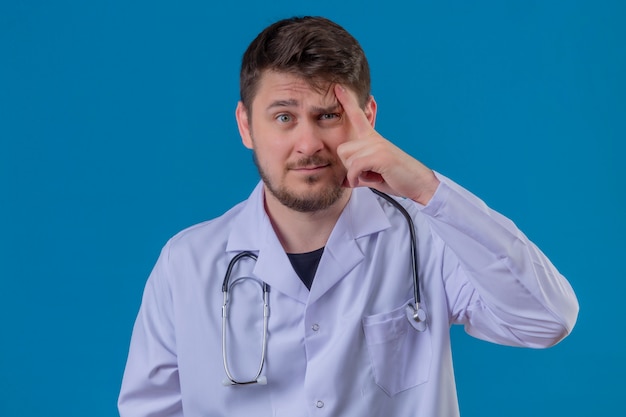 Young man doctor wearing white coat and stethoscope  having doubts pointing finger to head over isolated blue background