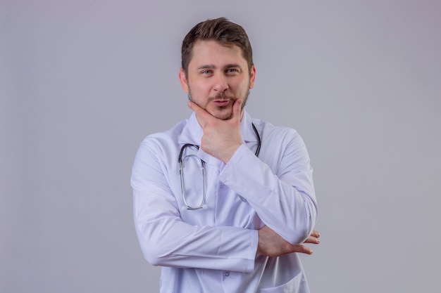 Free photo young man doctor wearing white coat and stethoscope  keeps hand under chin, thinks about something pleasant