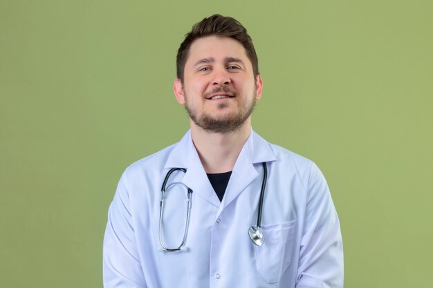 Young man doctor wearing white coat and stethoscope looking confident with smile on face over isolated green background