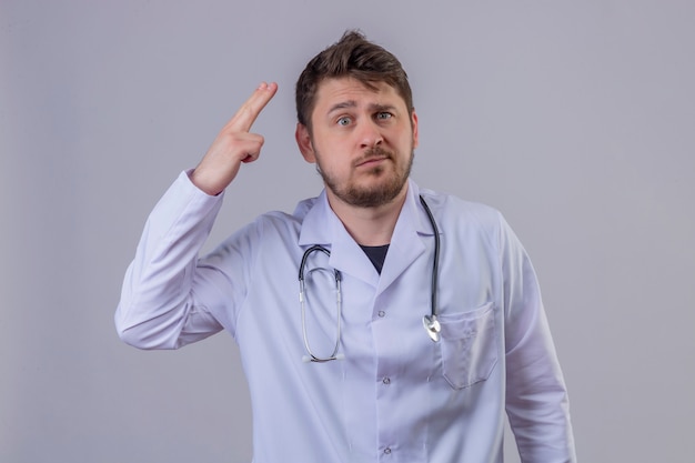 Young man doctor wearing white coat and stethoscope looking overworked doing gun gesture, suicide concept