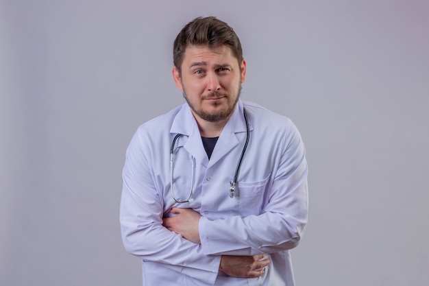 Young man doctor wearing white coat and stethoscope looking sick having strong pain in stomach standing