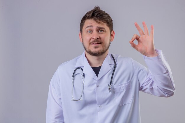 Young man doctor wearing white coat and stethoscope looking surprised and happy showing ok sign with his fingers