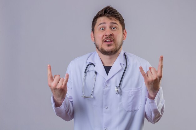 Young man doctor wearing white coat and stethoscope looking surprised and happy showing rock sign with his fingers