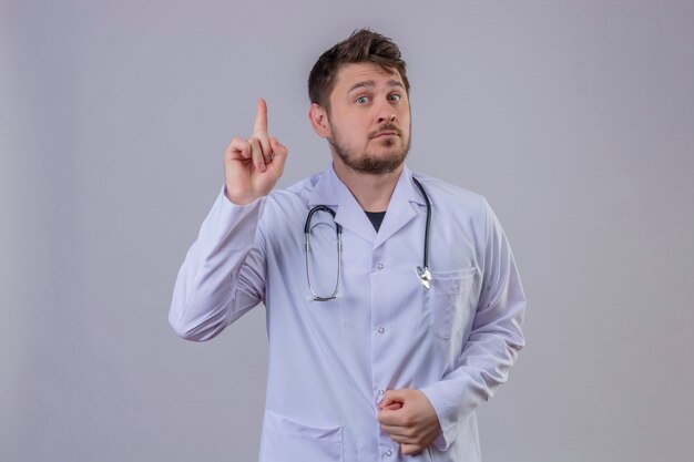 Young man doctor wearing white coat and stethoscope pointing finger up , new idea concept