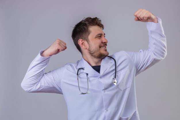 Young man doctor wearing white coat and stethoscope raising arms showing biceps expressing strength, winner concept