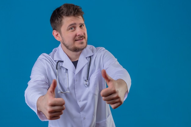 Доктор молодого человека нося белое пальто и стетоскоп показывая большие пальцы руки вверх показывать с счастливым лицом над изолированной голубой предпосылкой