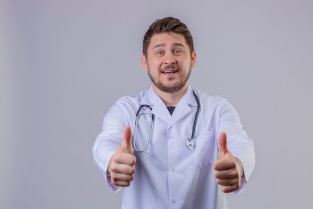 Free photo young man doctor wearing white coat and stethoscope showing thumbs up gesture with happy face