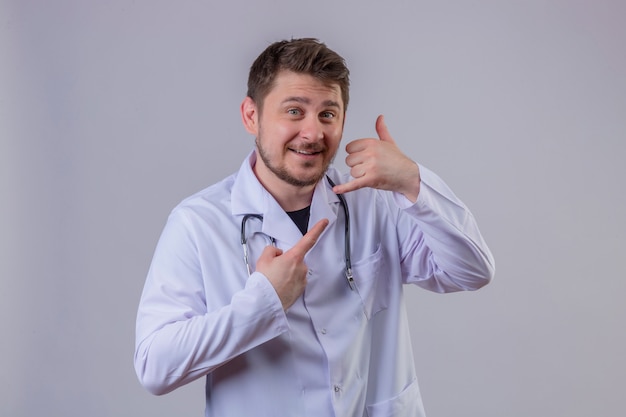 Young man doctor wearing white coat and stethoscope smiling doing talking on the telephone gesture call me sign and pointing to it