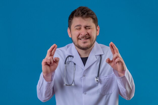 Young man doctor wearing white coat and stethoscope standing with closed eyes raising fingers crossed makes desirable wish over isolated blue background