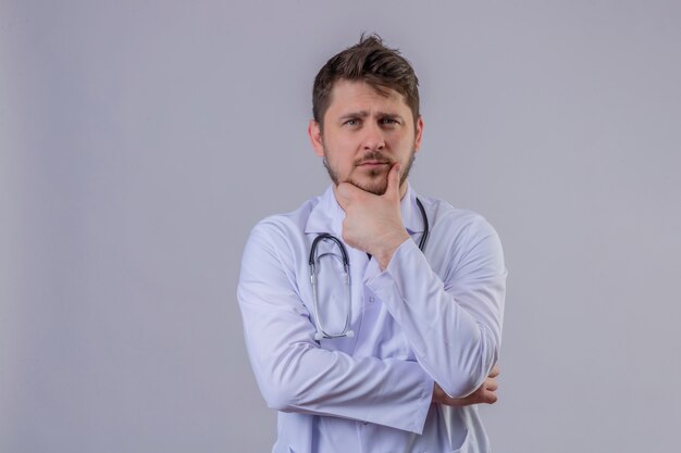 Young man doctor wearing white coat and stethoscope standing with skeptic facial expression touching his chin and thinking