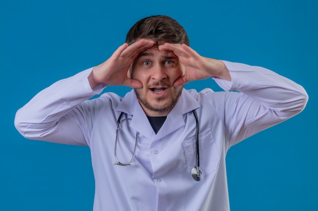 Young man doctor wearing white coat and stethoscope surprised looking far away with hand over head over isolated blue background