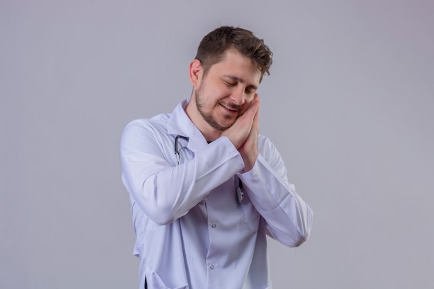 Young man doctor wearing white coat and stethoscope  tiered standing with hands together while smiling with closed eyes