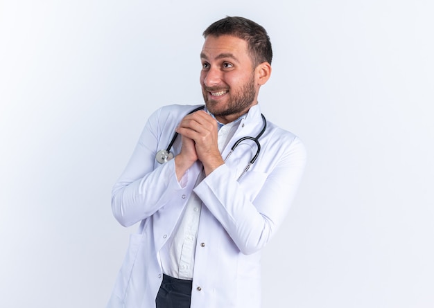 Young man doctor in white coat and with stethoscope around neck holding hands together happy and cheerful waiting for something