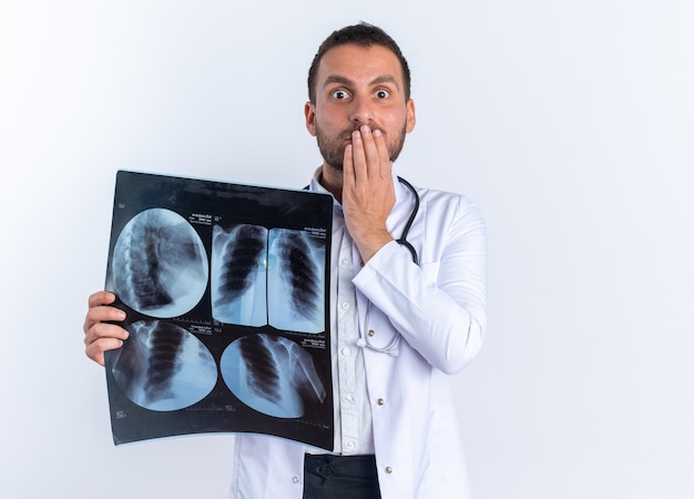 Young man doctor in white coat and with stethoscope around neck holding x-ray being shocked covering mouth with hand standing over white wall