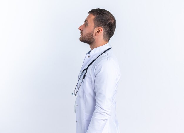 Young man doctor in white coat and with stethoscope around neck looking confident standing sideways over white wall