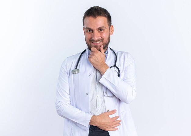 Молодой человек-врач в белом халате и со стетоскопом на шее выглядит счастливым и уверенным, уверенным в себе, улыбаясь.