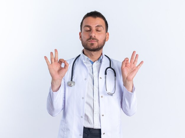 Молодой человек-врач в белом халате и со стетоскопом на шее расслабляет, делая жест медитации с пальцами, стоящими над белой стеной