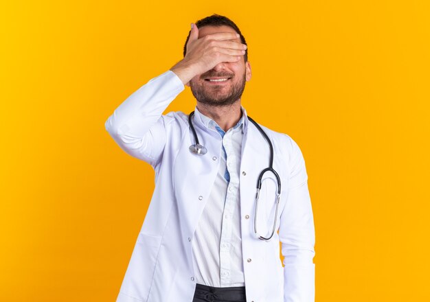 Young man doctor in white coat and with stethoscope around neck smiling cheerfully coning eyes with hand standing on orange