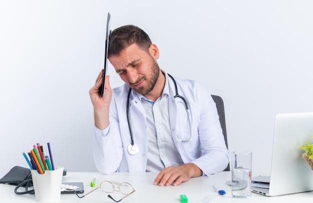 Young man doctor in white coat and with stethoscope holding clipboard over his head looking tired and overworked sitting at the table with laptop over white wall