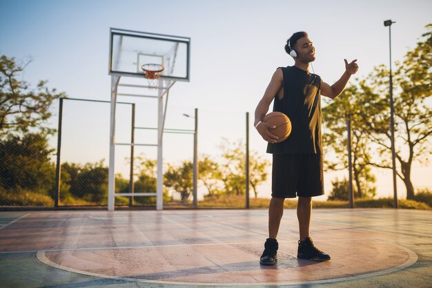 Young man doing sports, playing basketball on sunrise, listening to music on headphones