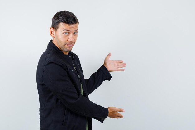 Young man doing welcome gesture in t-shirt, jacket and looking gentle. front view.