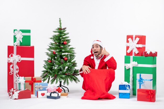 Молодой человек в костюме Санта-Клауса с подарками и украшенной елкой сидит в земле и делает жест «зови меня»