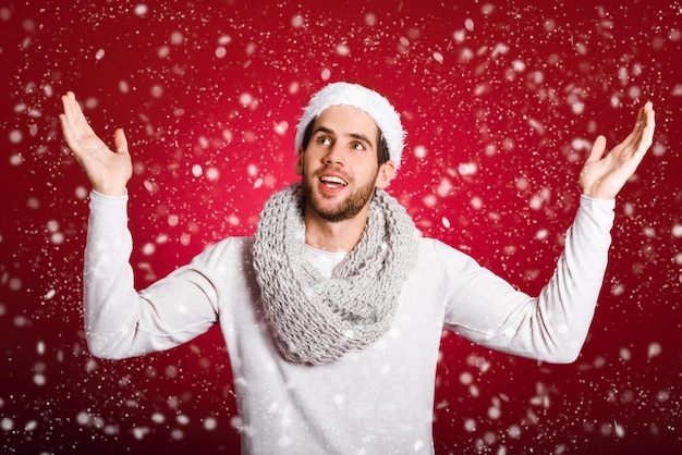 Молодой человек, одетый в зимнюю одежду под снегом на красном фоне парень с распростертыми объятиями и шляпу Санта Клауса