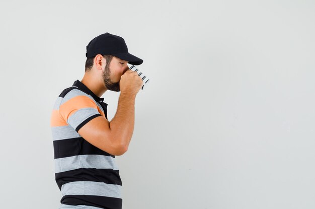 Young man drinking aromatic coffee in t-shirt, cap and looking pensive .