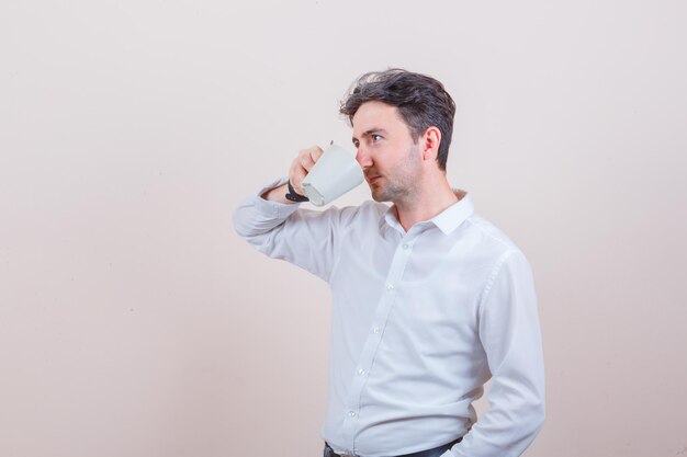 Young man drinking aromatic tea in white shirt and looking pensive