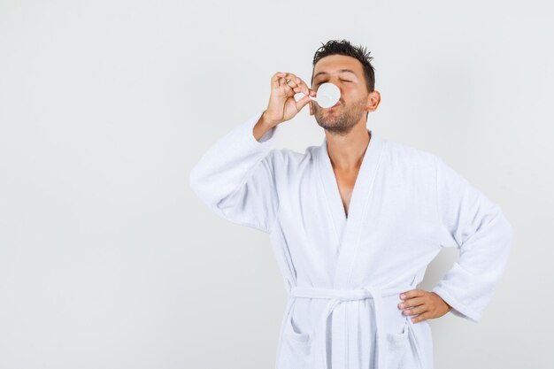 Young man drinking coffee after bath in white bathrobe front view.