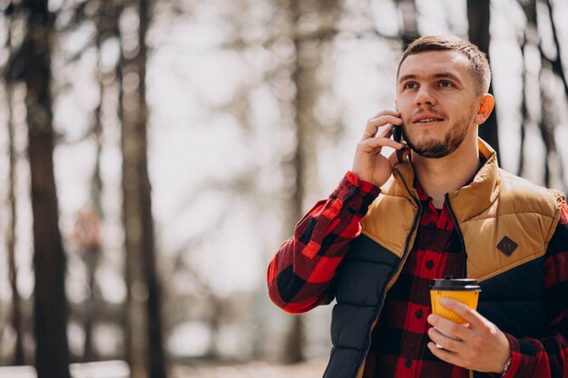 Молодой человек пьет кофе в парке и с помощью телефона