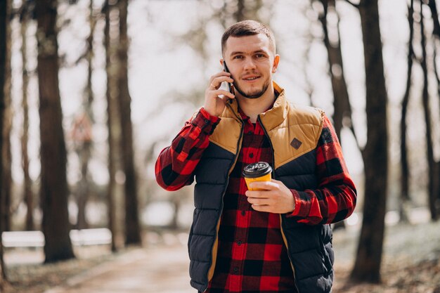 Молодой человек пьет кофе в парке и с помощью телефона