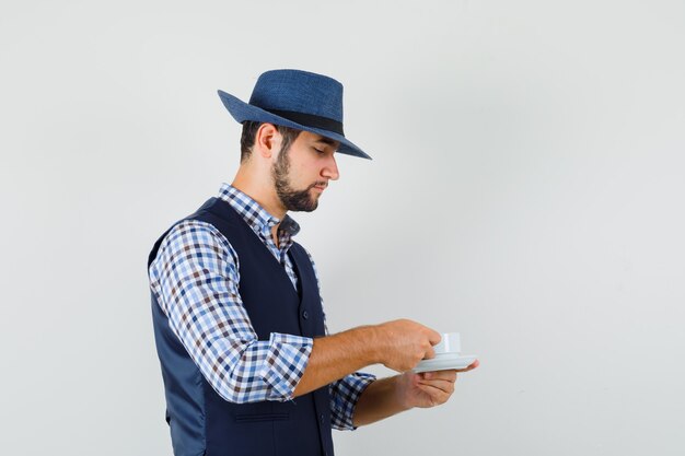 シャツ、ベスト、帽子でお茶を飲み、注意深く見ている若い男。 。