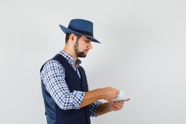 Young man drinking tea in shirt, vest, hat and looking careful. .