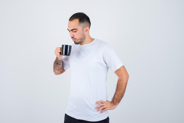 Молодой человек пьет чай, держа руку на талии в белой футболке и черных штанах и выглядит серьезным
