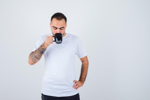 Young man drinking tea while holding hand on waist in white t-shirt and black pants and looking serious