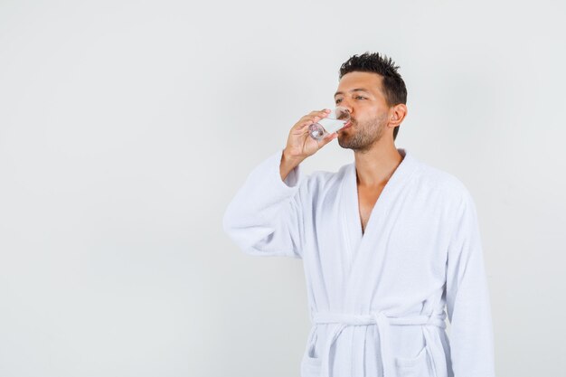 Young man drinking water in white bathrobe , front view.