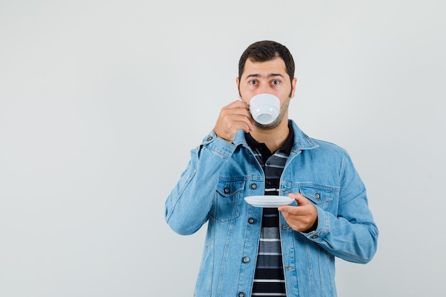 Young man drinknig tea in t-shirt, jacket and looking pensive. 