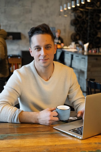 Free photo young man drinks tea and works on a laptop, freelancer working in a cafe
