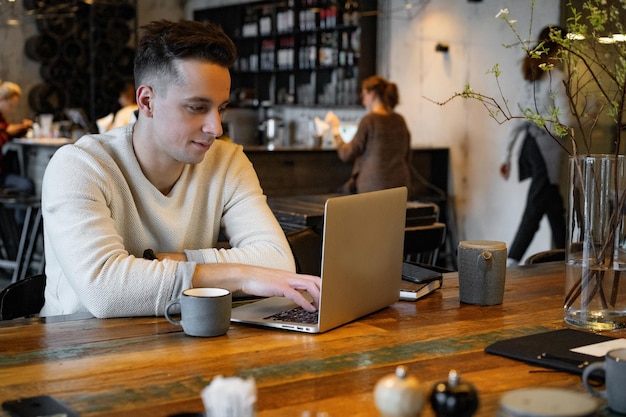 young man drinks tea and works on a laptop, freelancer working in a cafe
