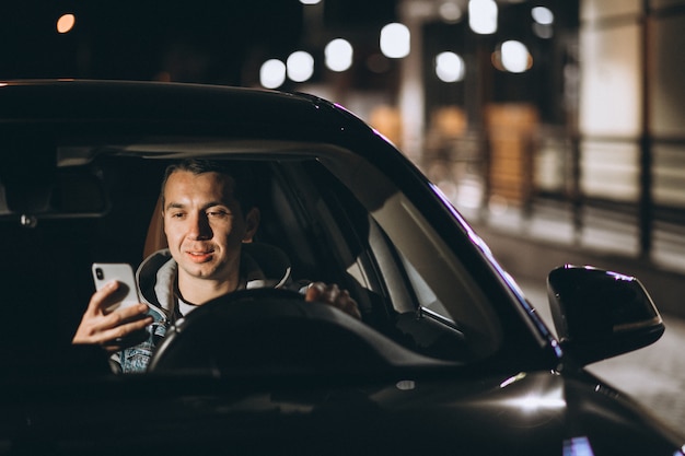 Молодой человек за рулем своего автомобиля в ночное время и разговаривает по телефону