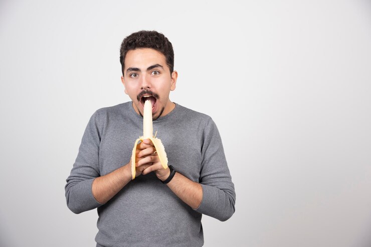 मुफ़्त फोटो a young man eating a banana over a white wall.
