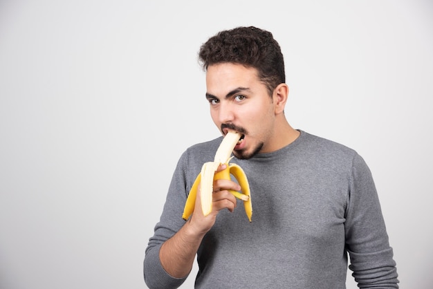 Молодой человек ест банан над белой стеной.