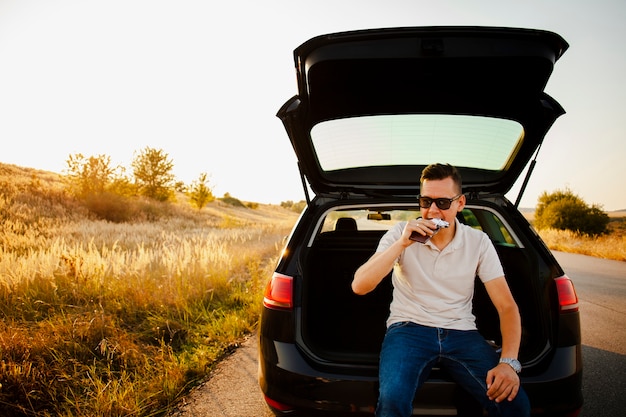 車のトランクの上に座ってチョコレートバーを食べる若い男