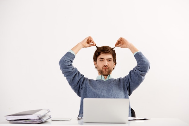 Young man employee working with laptop at office desk, grimacing and playing hair, procrastinating