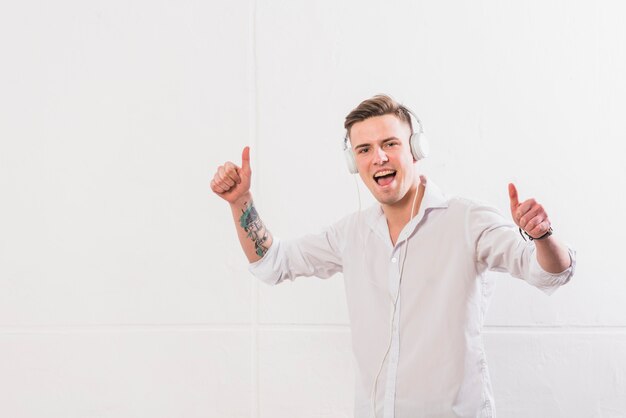 흰 벽에 가입 엄지 손가락을 보여주는 헤드폰에서 음악을 즐기는 젊은 남자